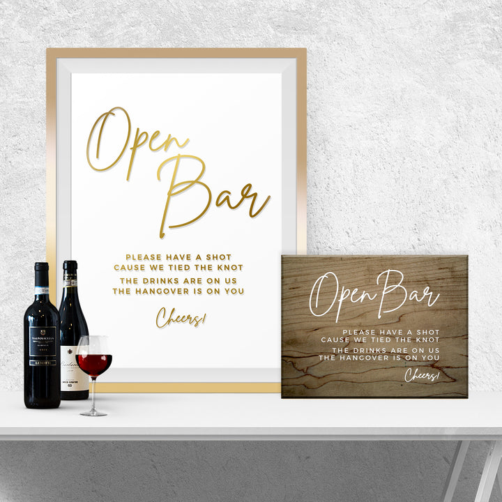 a framed sign with a message about an open bar, a wooden sign, and two bottles of wine on a white table.