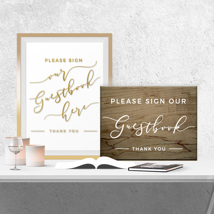 two signs on a table, one with a white background and gold lettering, and the other with a wooden background and white lettering. The signs ask guests to sign their guestbook, and there is a book and a candle on the table as well.