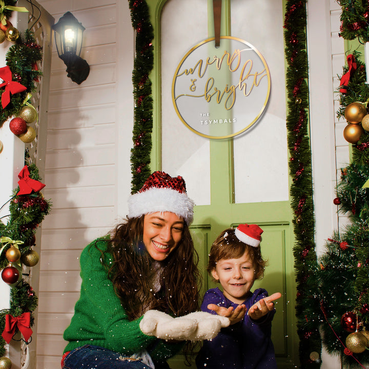 A woman and a child are standing in front of a Christmas-themed building, with the woman wearing a green sweater and a red and white hat, and the child wearing a red hat and smiling.