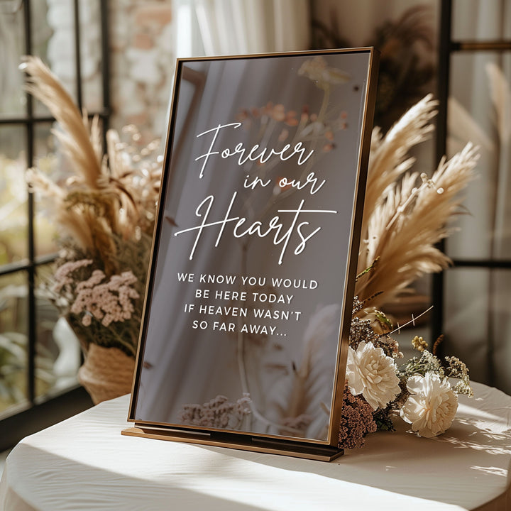 A sign with the message "Forever in our hearts" written on it, placed on a table with flowers and a window in the background.