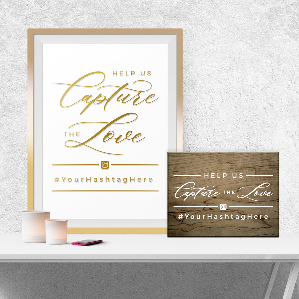 a framed sign with the text "Help us Capture the Love" and a smaller wooden sign with the same text. There is also a smartphone and a candle on a white surface.