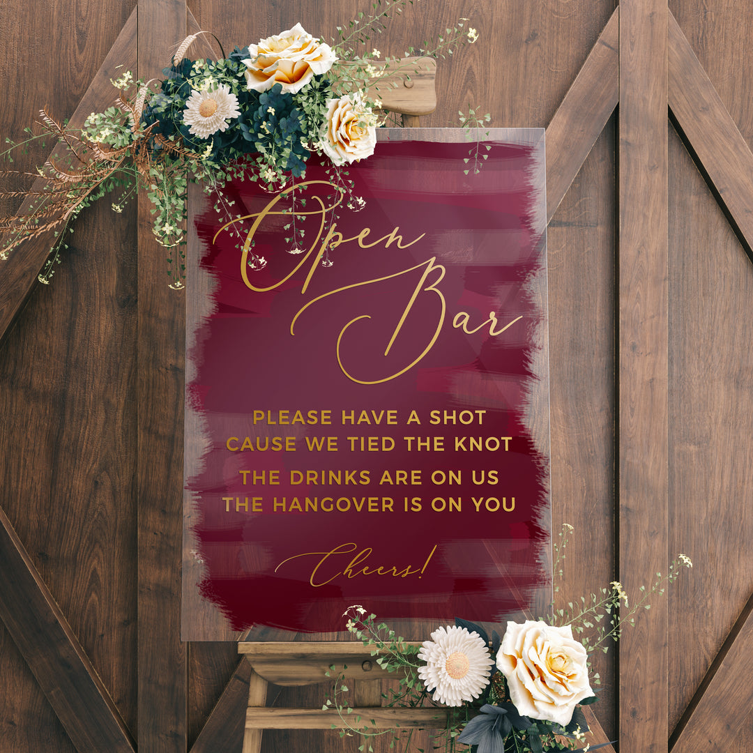 a sign with a maroon background and gold lettering, accompanied by a floral arrangement on a wooden stand.