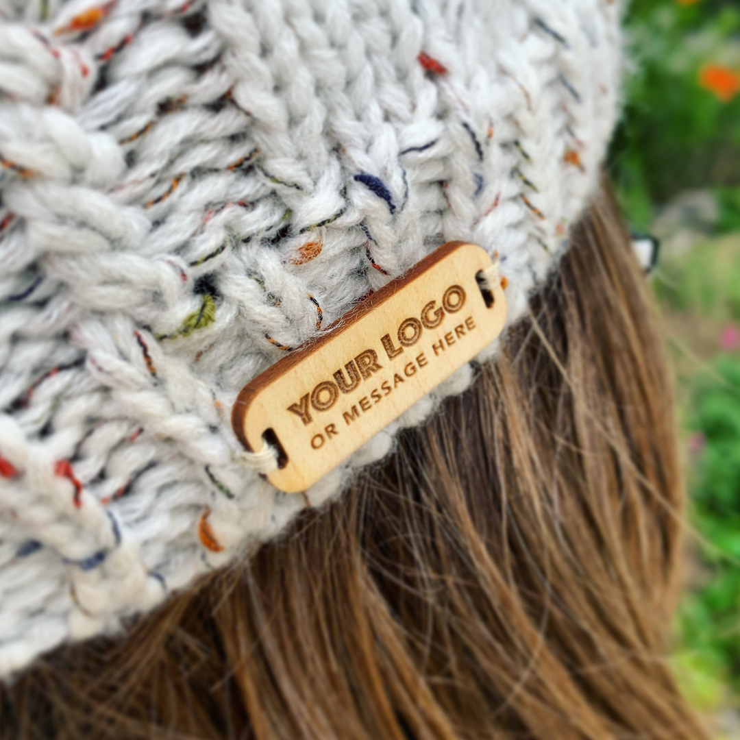 A close-up of a knitted hat with a wooden tag attached, displaying the text "YOUR LOGO OR MESSAGE HERE".