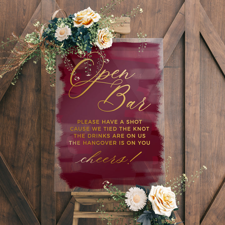 a sign with a maroon background and gold lettering, accompanied by a floral arrangement on a wooden stand.