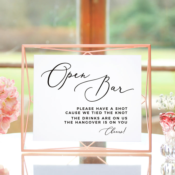 a sign that reads "Open Bar" in a pink frame, with a message that says "Please have a shot cause we tied the knot the drinks are on us the hangover is on you" in black text.
