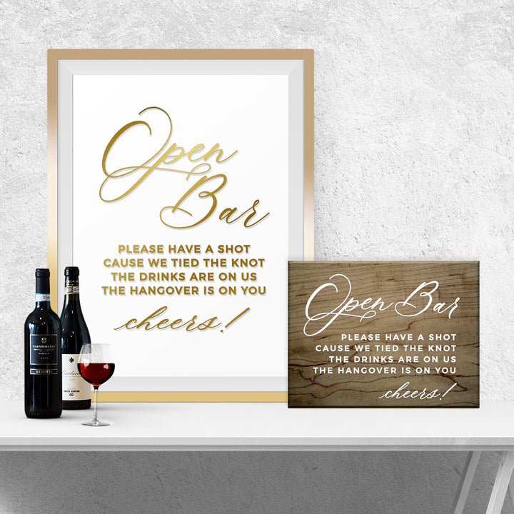 a framed sign with a message about an open bar, a wooden sign, and a wine bottle and glass on a white table.