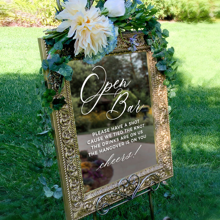 a decorative sign with a floral arrangement and greenery, placed in a grassy area. The sign has a message that reads "Open Bar" and includes a humorous instruction to tie the knot of the drinking vessel.
