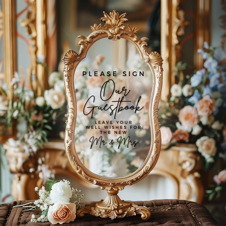 a decorative sign with a floral design, placed on a table with a vase of flowers. The sign reads "Please Sign Our Guestbook Leave Your Well Wishes for the New Mr & Mrs".
