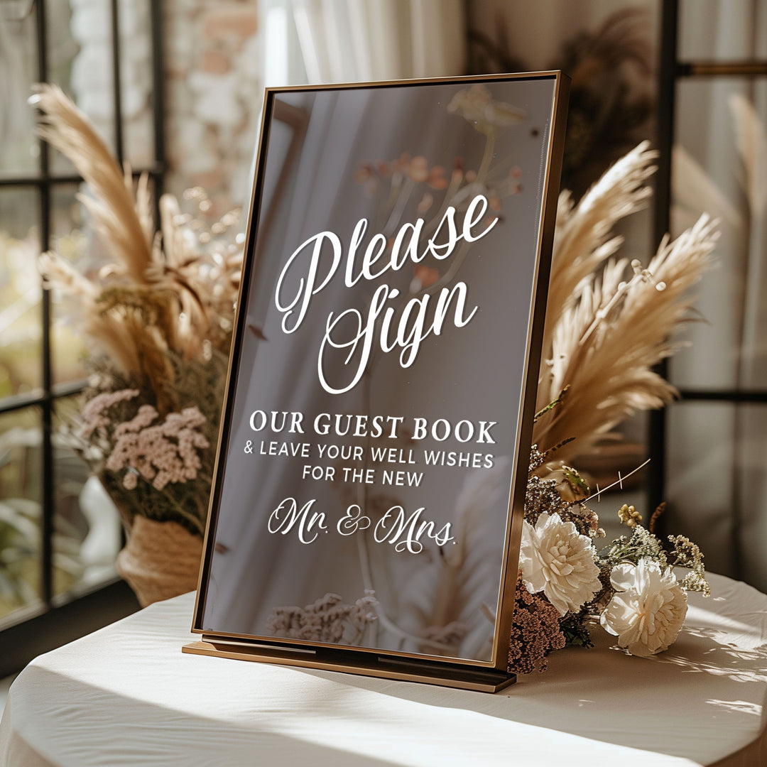 A rectangular sign with the text "Please Sign" and "OUR GUEST BOOK & LEAVE YOUR WELL WISHES FOR THE NEW Mr & Mrs" is displayed on a table with flowers in the background.
