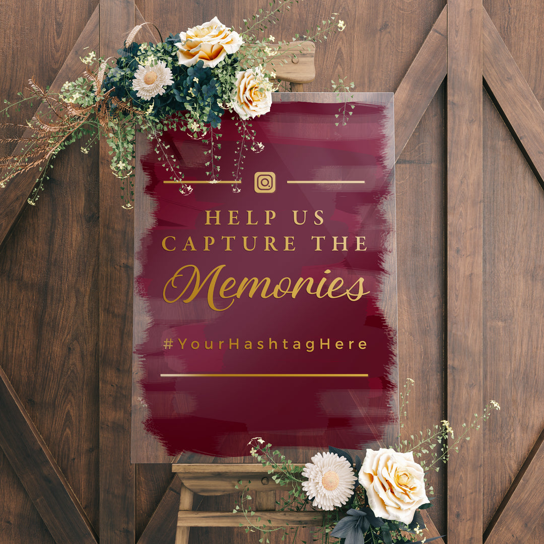 a sign with a maroon background and gold text, placed on a wooden easel. The sign reads "Help us capture the memories" and includes a hashtag "#YourHastagHere".