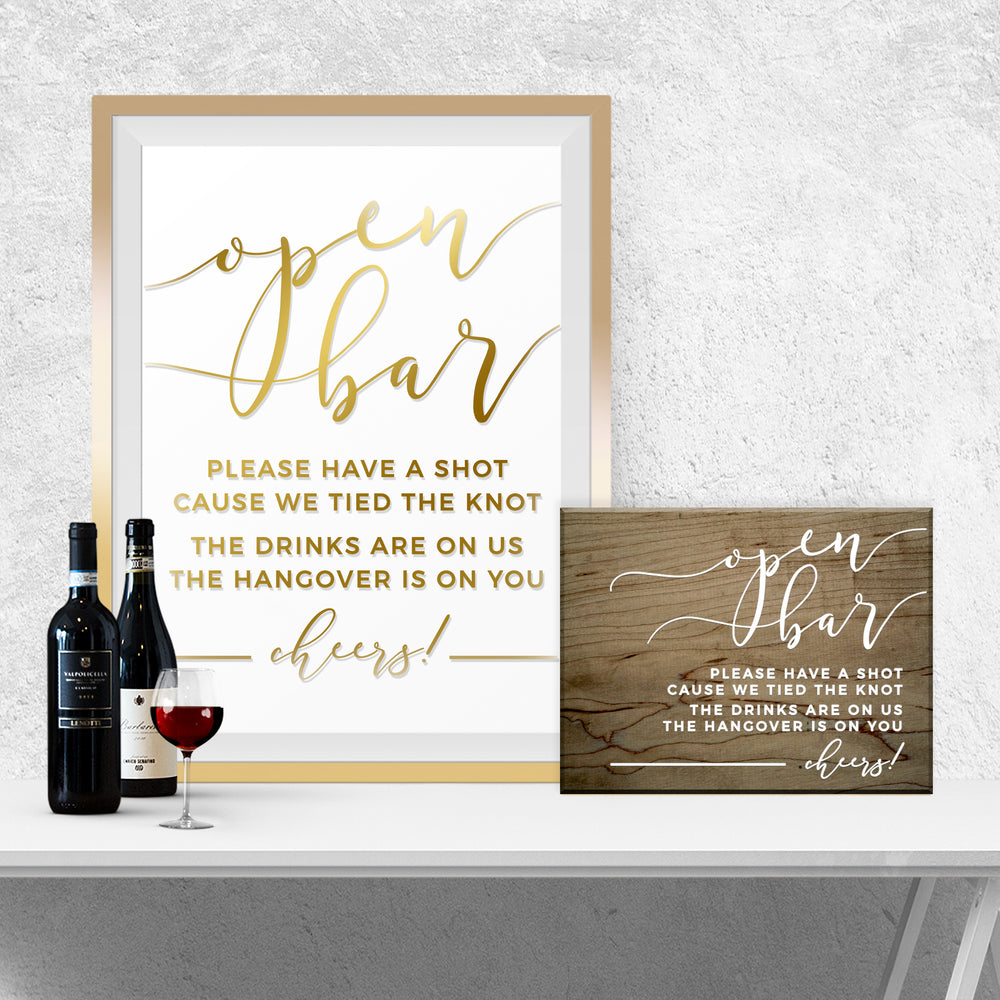 a framed sign with a message about an open bar, a wooden sign, and two bottles of wine on a white table.