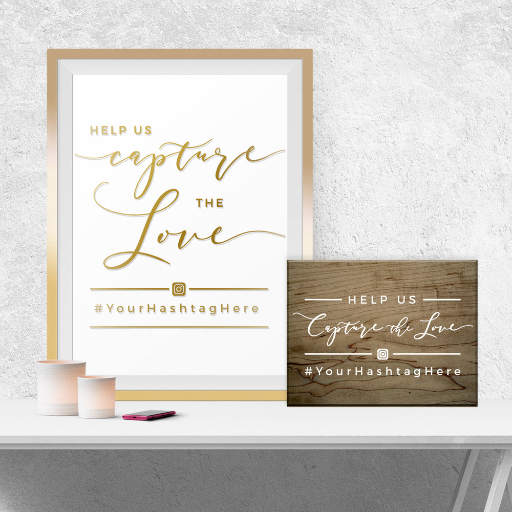 a framed sign with the text "Help us capture the love" and a smaller wooden sign with the same text. There is also a smartphone and a candle on a white surface.
