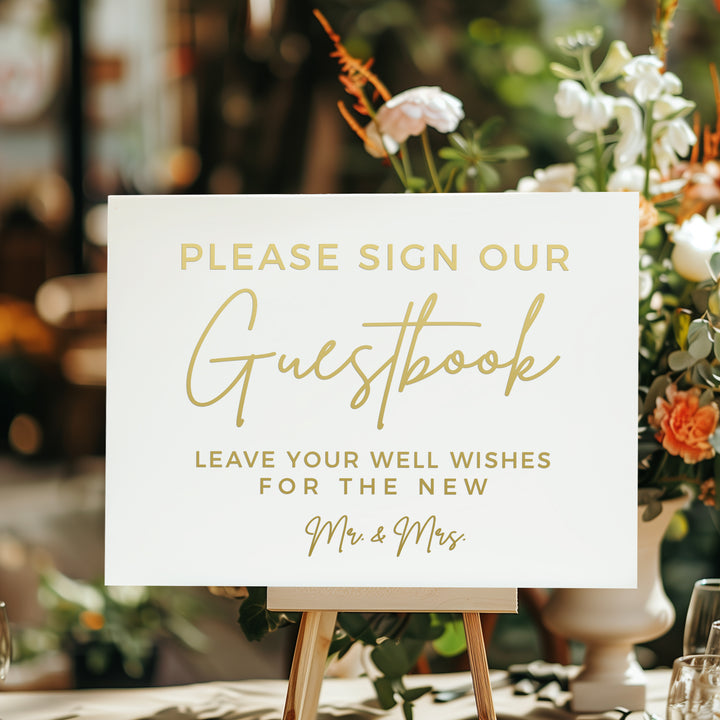 a sign that reads "Please sign our guestbook. Leave your well wishes for the new Mr & Mrs." The sign is placed on a table with a vase of flowers in the background.