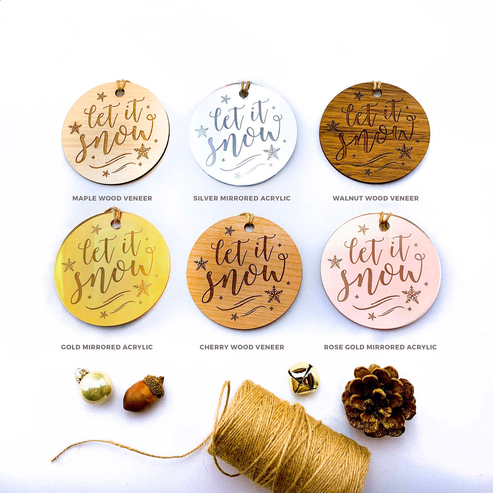 a collection of six round ornaments with the phrase "let it snow" engraved on them, arranged on a white background. The ornaments come in various colors and materials, including wood veneer, silver acrylic, walnut wood veneer, gold mirrored acrylic, cherry wood veneer, and rose gold mirrored acrylic. There are also some ornaments and a spool of twine or string present in the image.