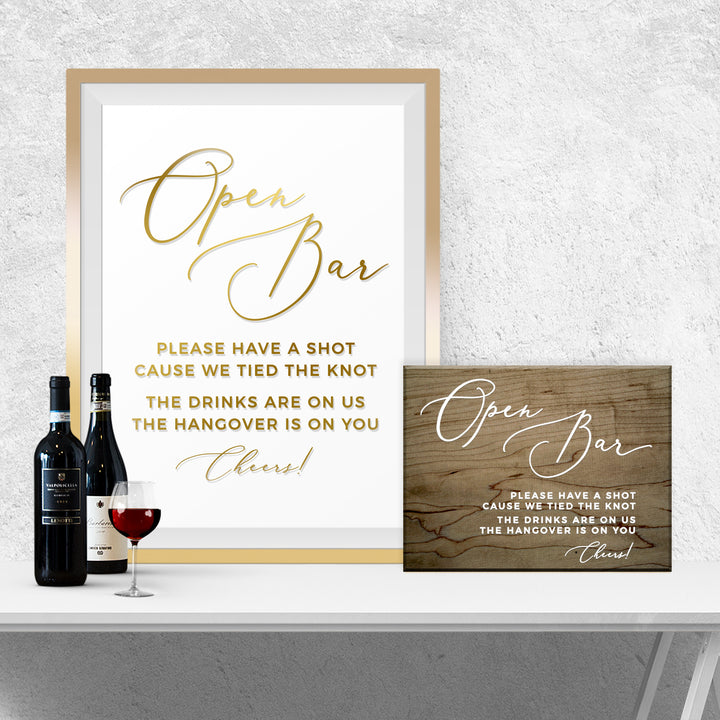 a framed sign with a message about an open bar, a wooden sign, and two bottles of wine on a white table.