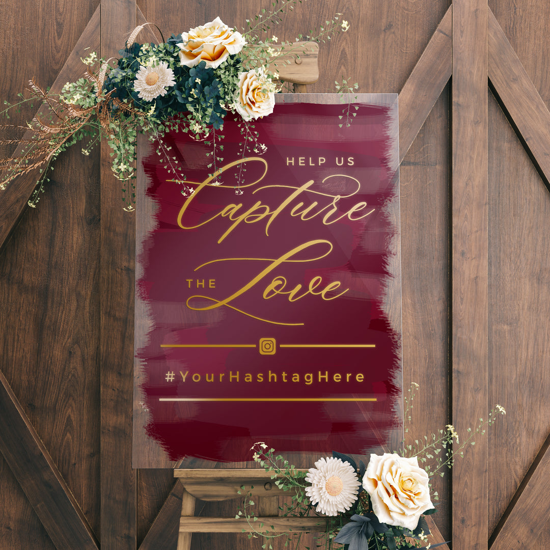 a sign with a maroon background and gold cursive text, which reads "Help us Capture the Love" and "#Your HashtagHere". The sign is surrounded by flowers and greenery, and there is a wooden easel in the background.
