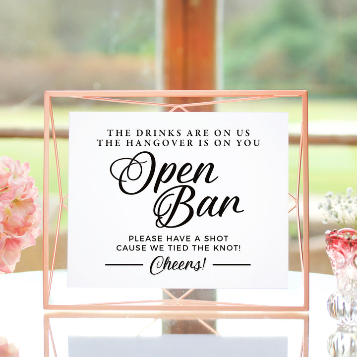 a sign that reads "The drinks are on us, the hangover is on you. Open Bar. Please have a shot. Cause we tied the knot! Cheers!" The sign is placed on a table with a vase of flowers in the background.
