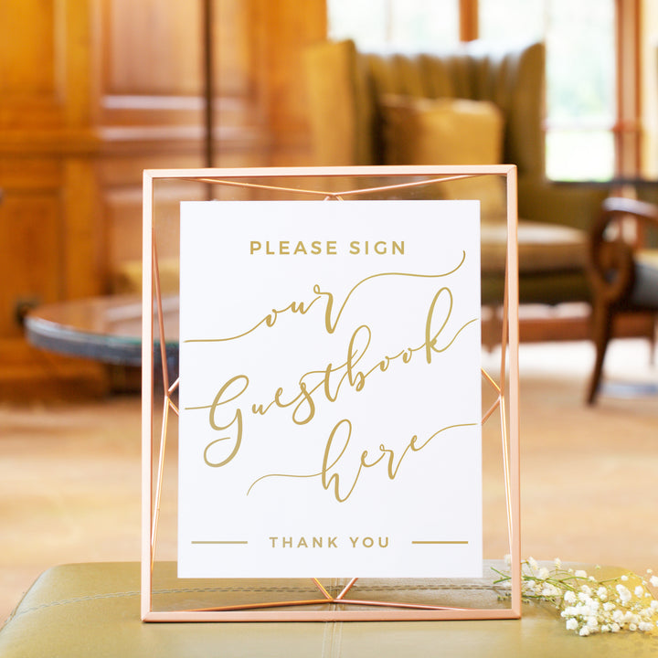 A sign that reads "Please Sign our Guestbook here" in a gold cursive font, placed on a table with a floral arrangement.