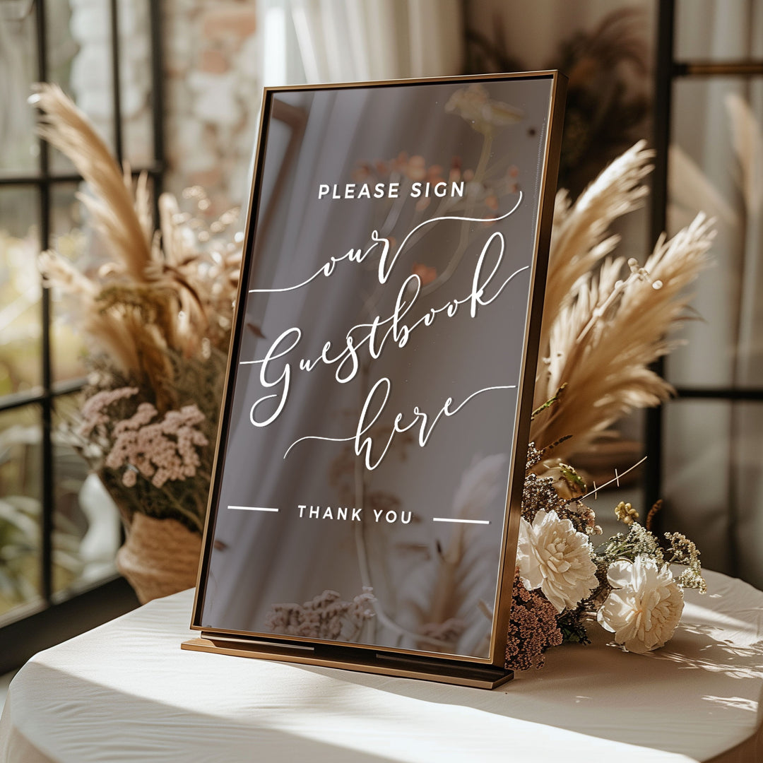 A sign that reads "Please Sign our Guestbook here" with a "Thank you" message below it, placed on a table with flowers.