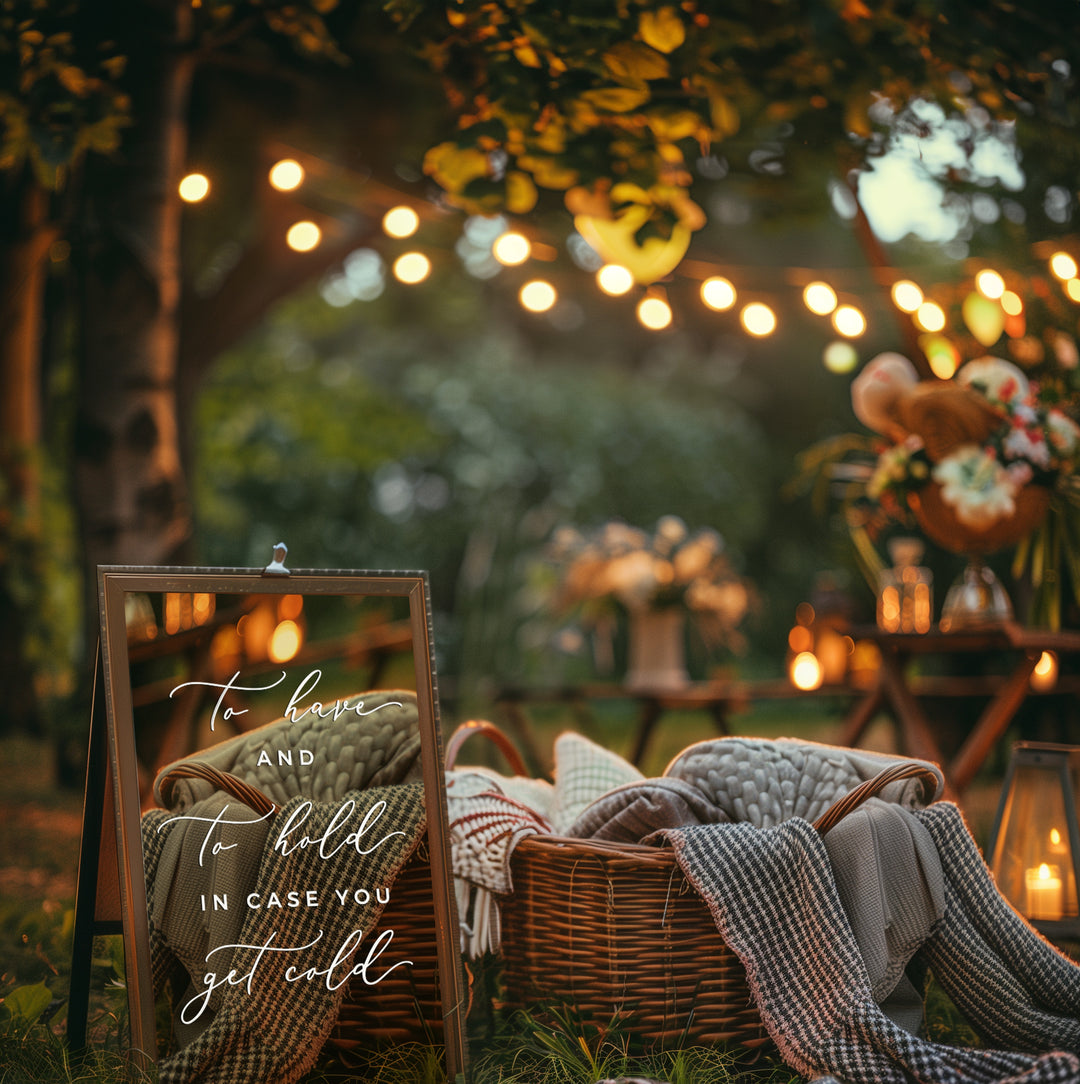 a cozy outdoor setting with a wicker basket filled with blankets and a sign that reads "To know and to hold in case you get cold". The scene is set in a garden or park, with string lights and lanterns creating a warm and inviting atmosphere.