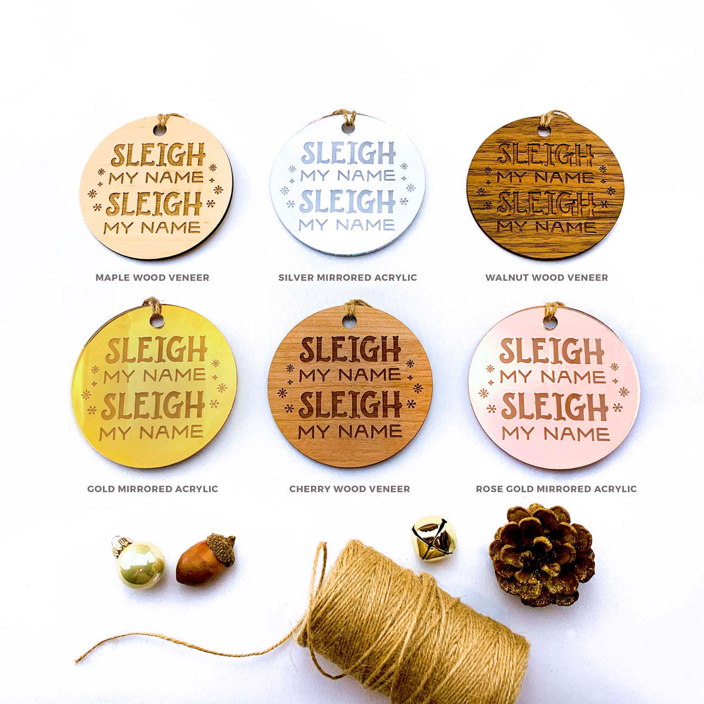 six circular ornaments with the text "SLEIGH MY NAME Sleigh MY NAME Sleigh MY NAME" printed on them, along with some natural elements like pine cones and acorns.