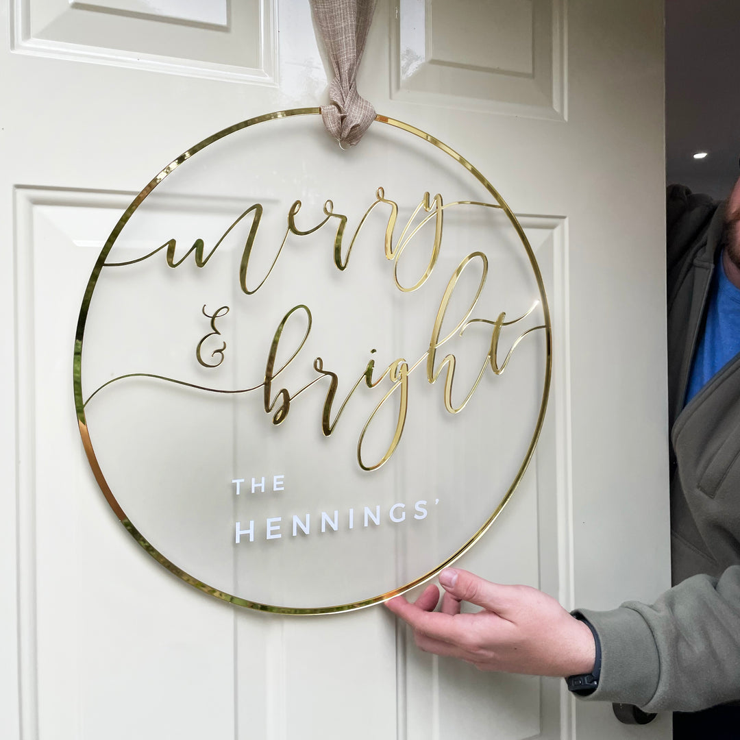 A person is holding a clear, gold-colored sign that says "Merry & Bright" in a festive style.