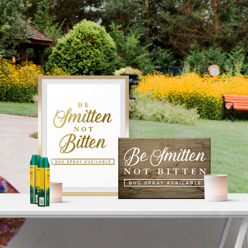 two signs on a table, one with a gold frame and the other with a wooden frame, both displaying the message "Be Smitten Not Bitten Bug Spray Available".