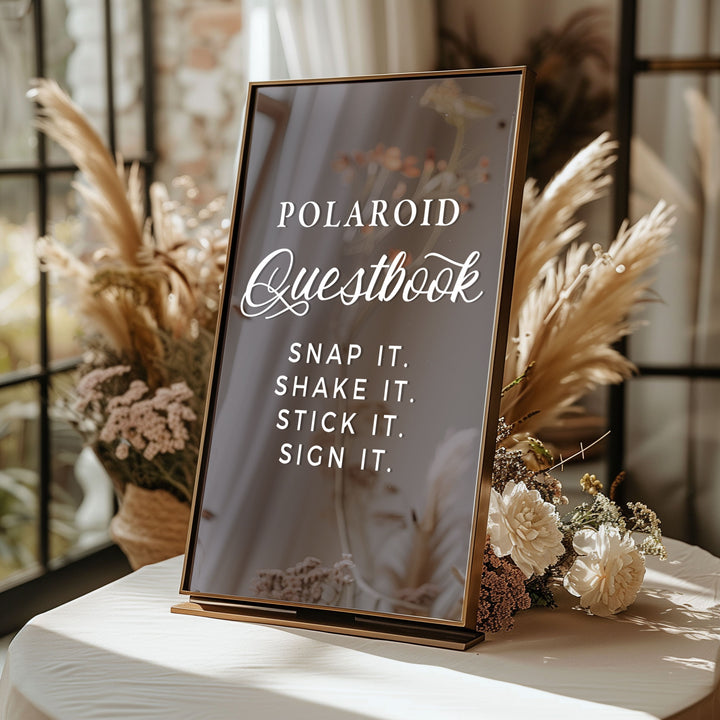 A black framed sign with white text that reads "POLAROID QUESTBOOK" and "SNAP IT. SHAKE IT. STICK IT. SIGN IT." is placed on a table with flowers in the background.