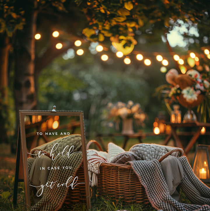 a cozy outdoor setting with a sign that reads "To have and to hold in case you get cold". The scene includes a wicker basket filled with blankets, a vase of flowers, and string lights creating a warm and inviting atmosphere.