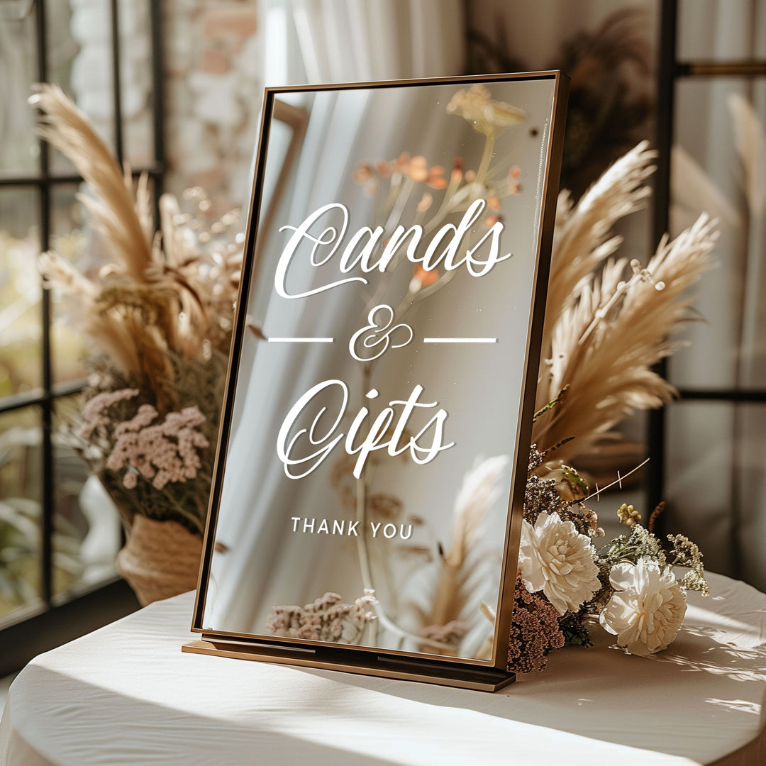 A mirror-like sign displaying the text "Cards & Gifts" and "Thank you" is placed on a table with flowers and pom poms.