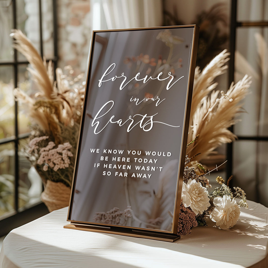 A black sign with white text that reads "Forever in our hearts" is displayed on a table, surrounded by flowers and a window.