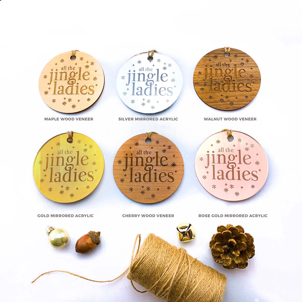 a collection of six circular ornaments with the text "all the jingle ladies" printed on them, each with a different color and material. The ornaments are arranged on a white background, along with some twine, a pine cone, and other small items.