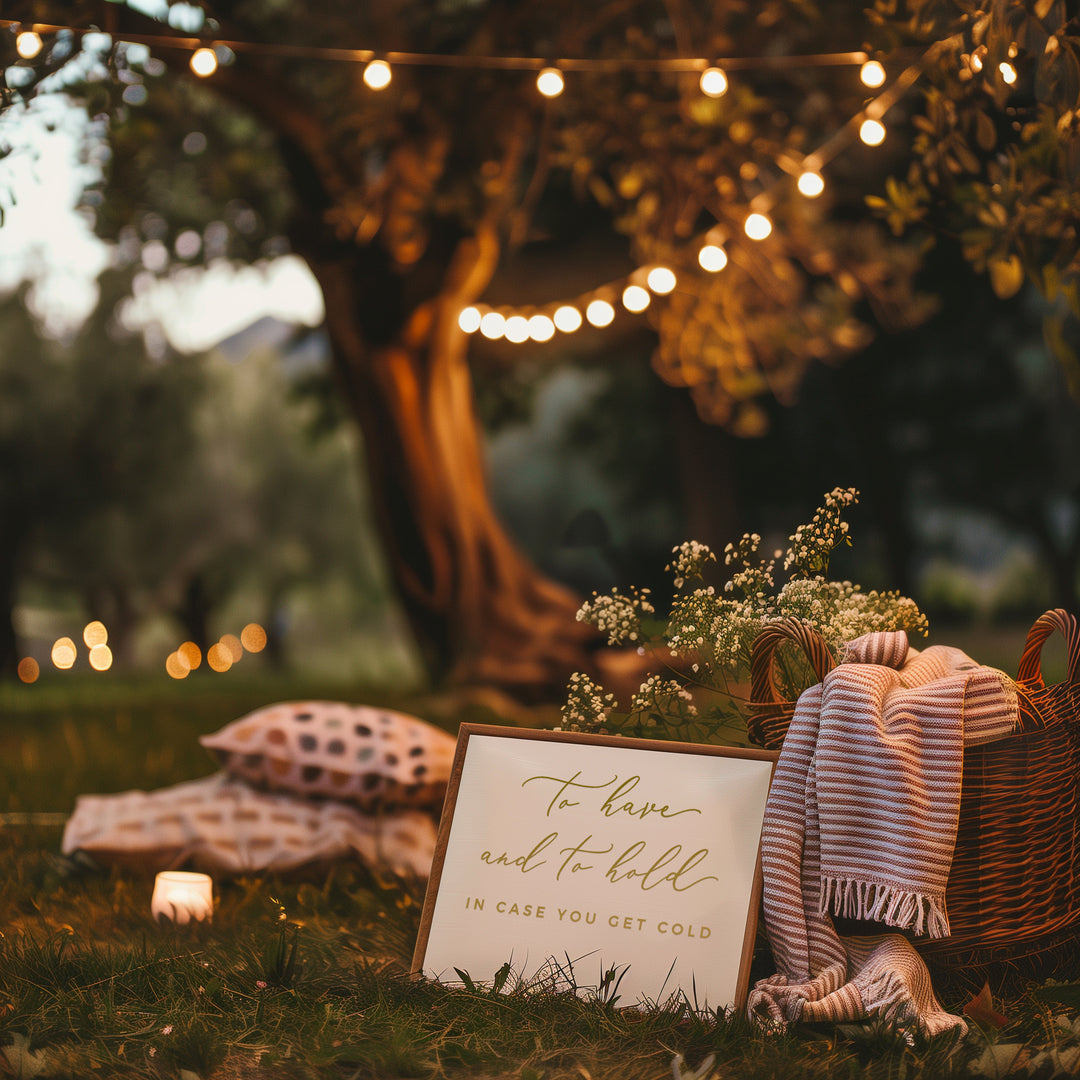 a cozy outdoor setting with a sign that reads "To have and to have in case you get cold", surrounded by a wicker basket, a blanket, and a candle. The scene is set against a backdrop of a tree with lights strung around it, creating a warm and inviting atmosphere.