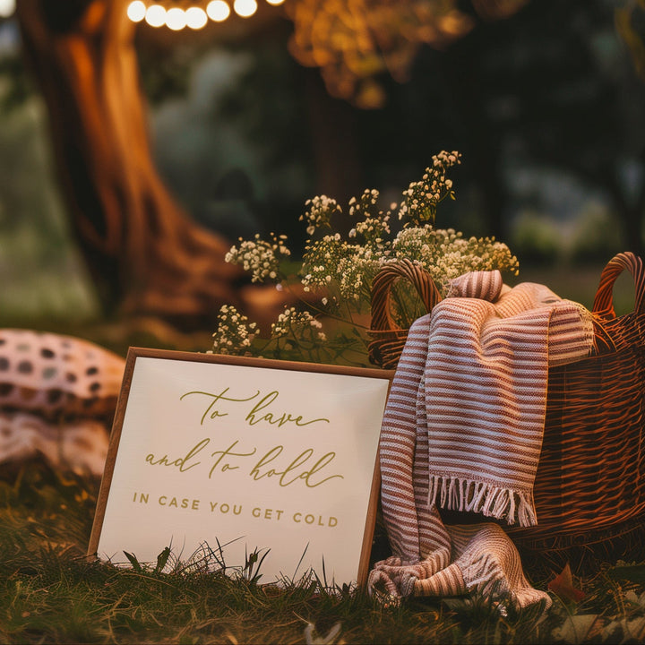 a cozy outdoor setting with a sign that reads "To have and to have in case you get cold", surrounded by a wicker basket, a blanket, and a candle. The scene is set against a backdrop of a tree with lights strung around it, creating a warm and inviting atmosphere.