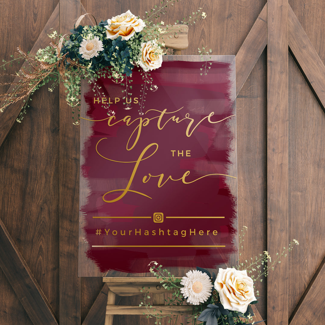 a sign with a maroon background and gold cursive text that reads "Help us capture the love" and "#Your HashtagHere". The sign is surrounded by flowers and greenery, and there is a wooden easel in the background.