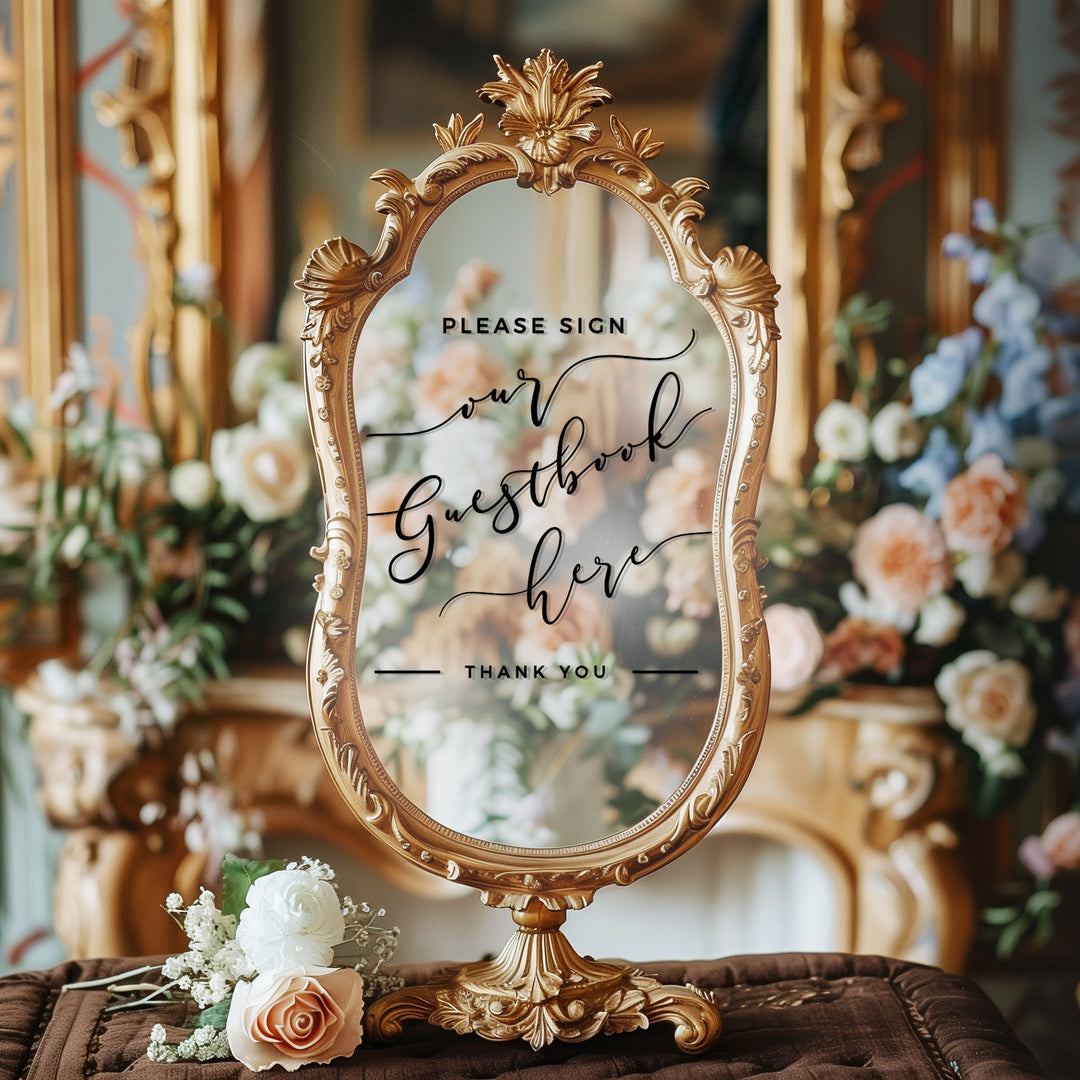 A decorative mirror with a sign in front of it, placed on a table with flowers.