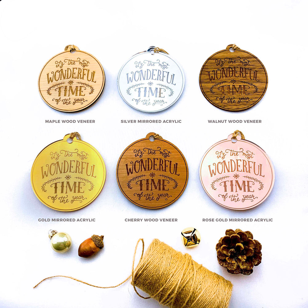 a collection of six round wooden ornaments with the phrase "It's the most wonderful time of the year" engraved on them, arranged in a grid with three rows and two columns. The ornaments come in various colors, including gold, silver, and wood veneer, and are accompanied by other decorative items such as a pine cone, a gold ornament, and a spool of twine.