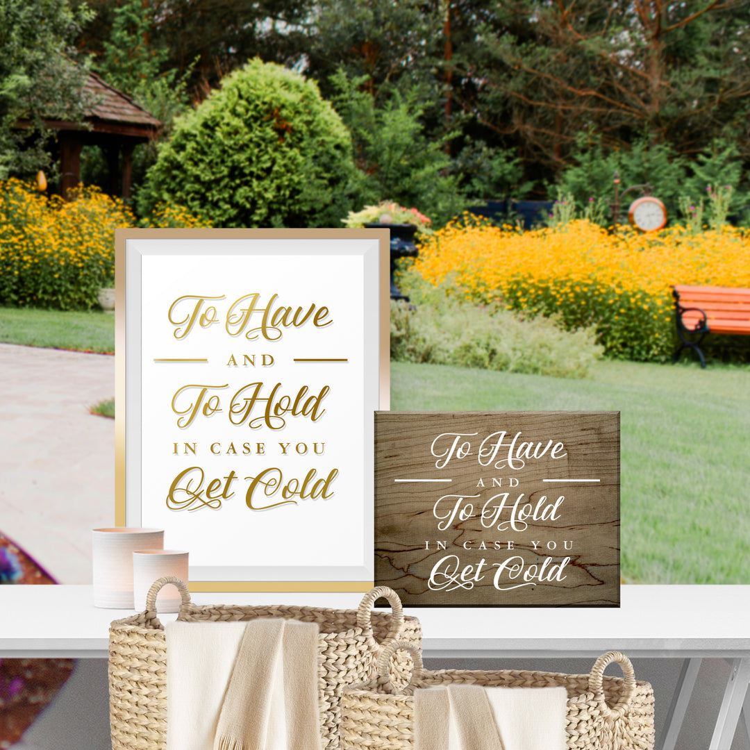 a framed sign with the text "To Have and To Hold in Case You Get Cold" and a wooden sign with the same text. The sign is placed on a table, and there are baskets nearby. The background shows a garden with yellow flowers and a bench.