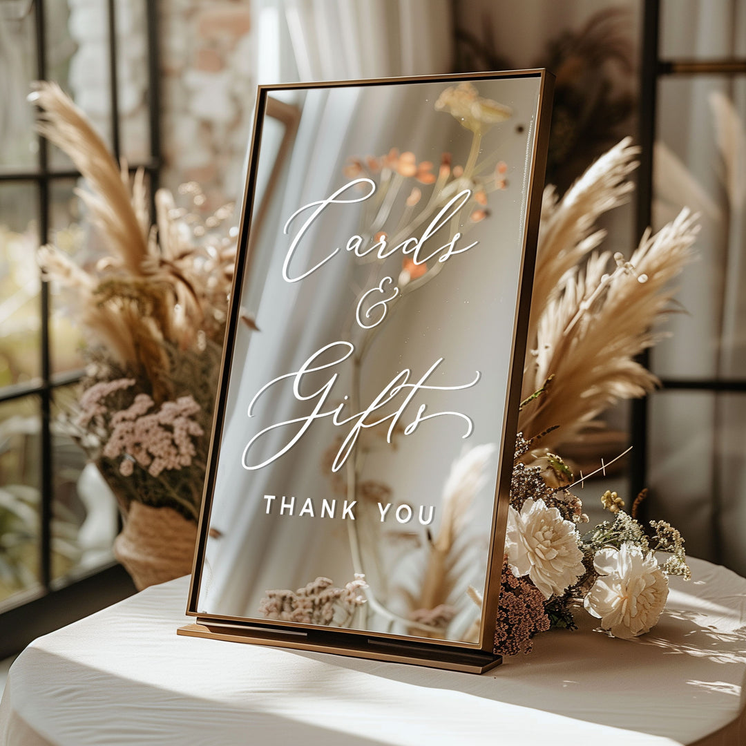 A mirror-like sign displaying the text "Cards & Gifts Thank You" is placed on a table with flowers and pom poms in the background.